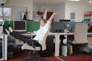 Side view of cheerful female employee in formal outfit sitting on office chair with crossed legs on desk and stretching