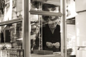 Black and white candid shot of a senior man at a kiosk in an urban setting, showcasing reflection and urban life.