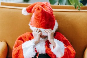 Little anonymous boy hiding face with bright red fluffy Santa hat while celebrating holiday in daytime
